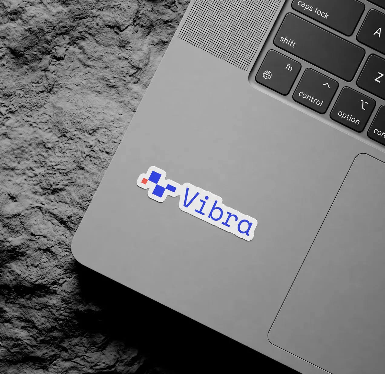 Un sticker de Vibra pegado en la tapa de una laptop, sobre un fondo de piedra